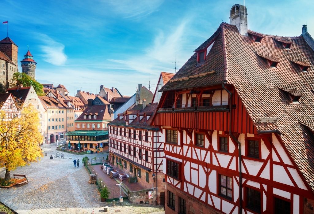 Halftimbered medieval houses with red wood and white walls on the right, a small city square in the center and a stone tower to the left