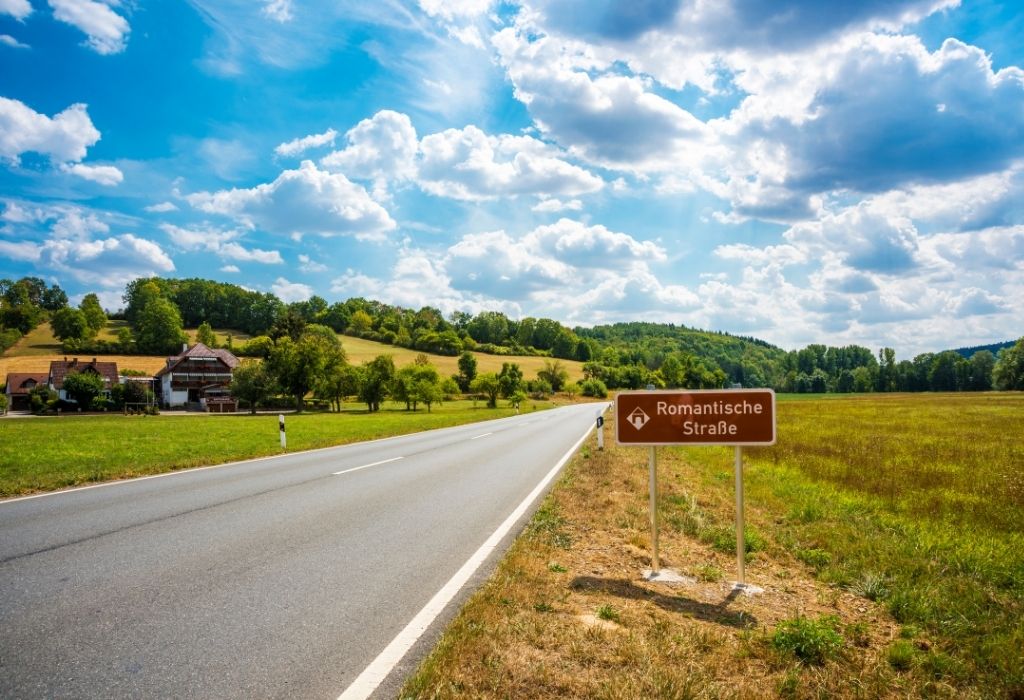 Small country road in Bavaria through a field and a small group of houses  with a brown sign on the right "Romanic Road"